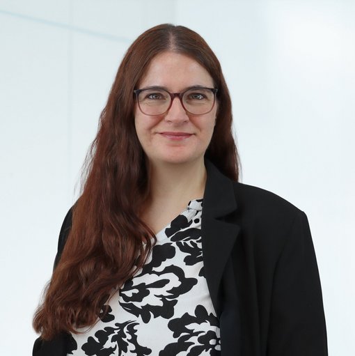 The picture shows a woman with long reddish-brown hair and glasses. She is wearing a black and white patterned blouse and a black blazer. She smiles friendly into the camera.