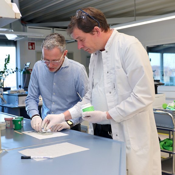 Auf dem Bild sind zwei Männer in einem Labor zu sehen, die mit Lotpaste experimentieren. Der Mann links hat graue Haare, trägt eine Brille und ein hellblaues emd. Der Mann rechts hat braune Haare und trägt einen weißen Kittel.