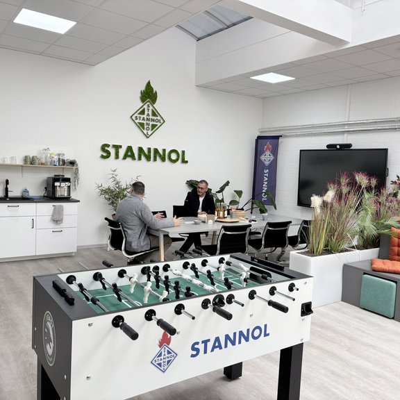 The picture shows a large training room with a table football table in the foreground and rows of seats with orange cushions on the right. In the background is a table with two people sitting at it. A white kitchenette can be seen at the back on the right and a large monitor on the left. A large Stannol logo made of moss hangs on the wall in the background. 