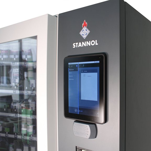 The picture shows a section of Stannol's Smart Warehouse: The touch display for operation can be seen in the foreground, with the Stannol logo above it. To the left, some stored products can be seen through the glass front.