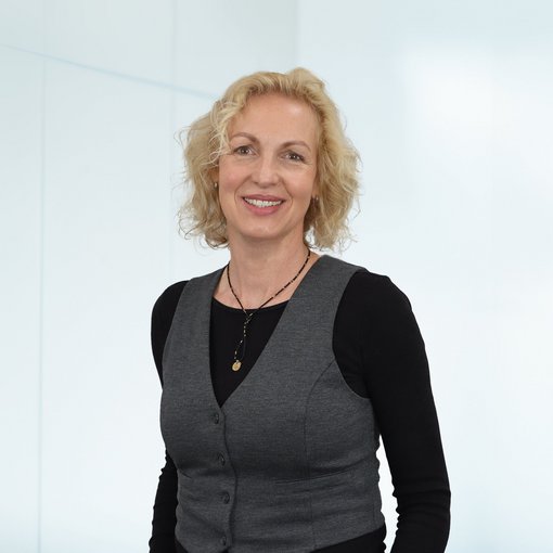 The picture shows a woman with light blonde, half-length wavy hair. She is wearing a black shirt, a grey waistcoat and a black and gold necklace. She smiles friendly into the camera.