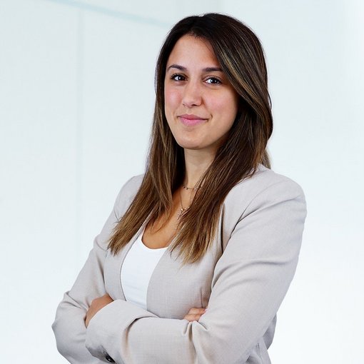 Auf dem Bild ist eine junge Frau mit langen braunen Haaren zu sehen. Sie trägt ein weißes Top und einen hellbeigen Blazer. Sie lächelt freundlich in die Kamera.